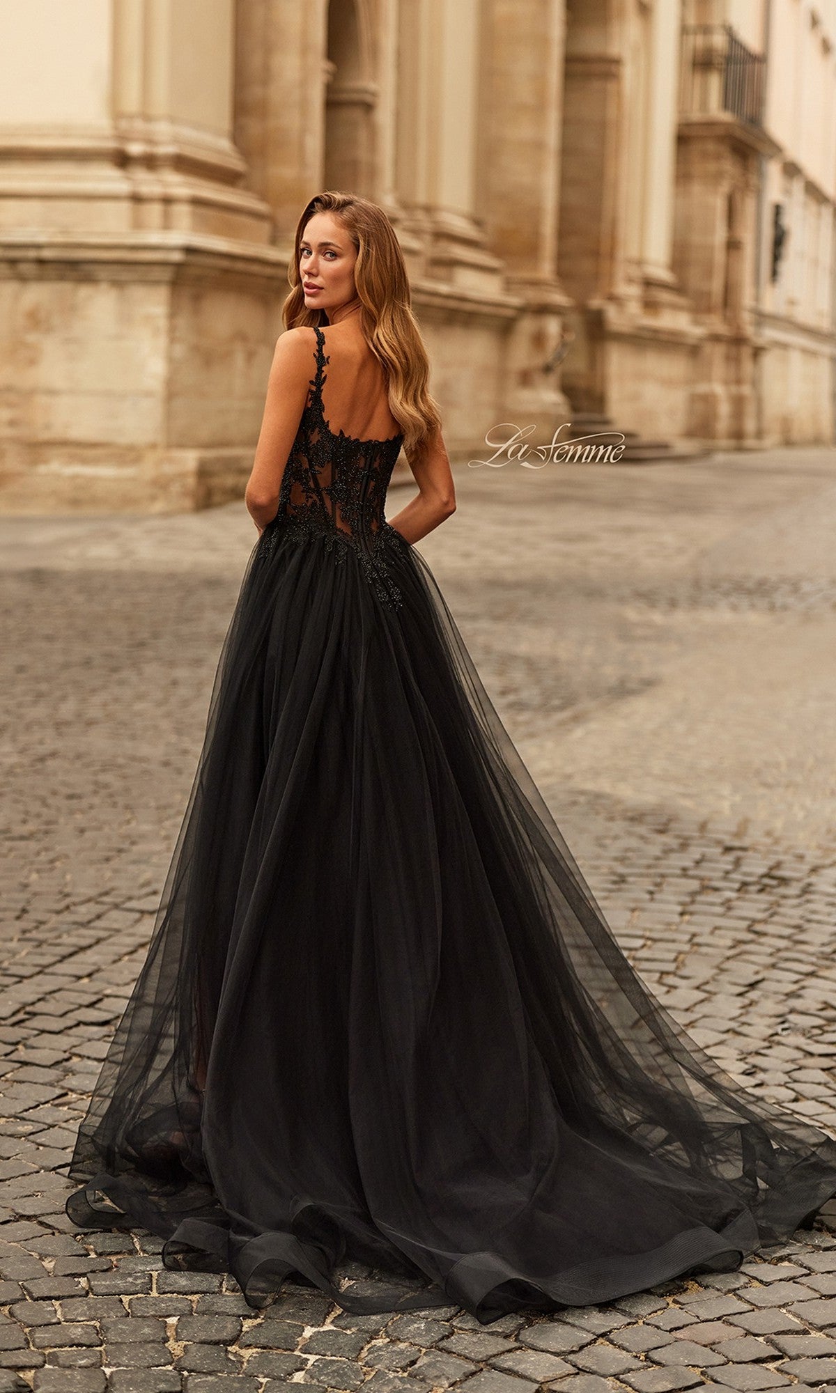 A woman in the La Femme 33499 long prom dress, featuring a black sheer floor-length skirt and fitted lace bodice with delicate details, stands on a cobblestone street, looking over her shoulder.